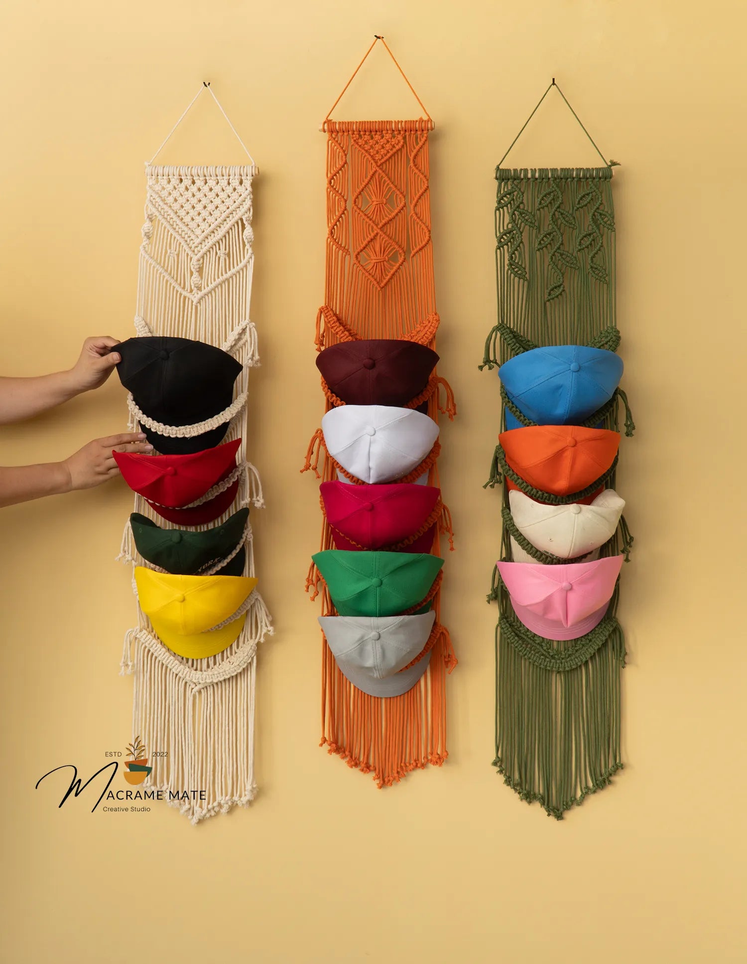 Three macrame hat racks in different colors with colorful hats on a yellow background.