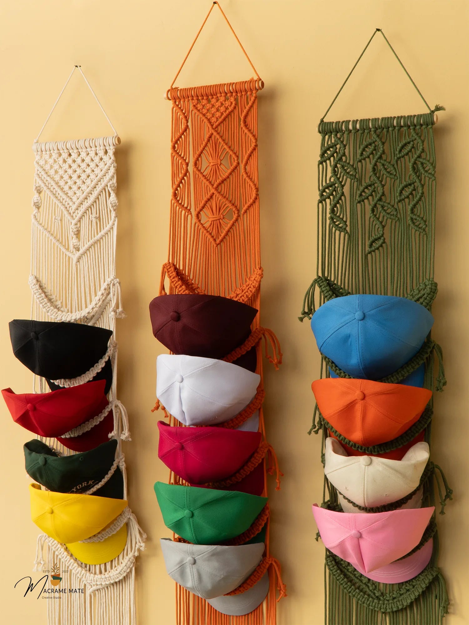 Three macrame hat racks in different colors hanging on a beige wall.