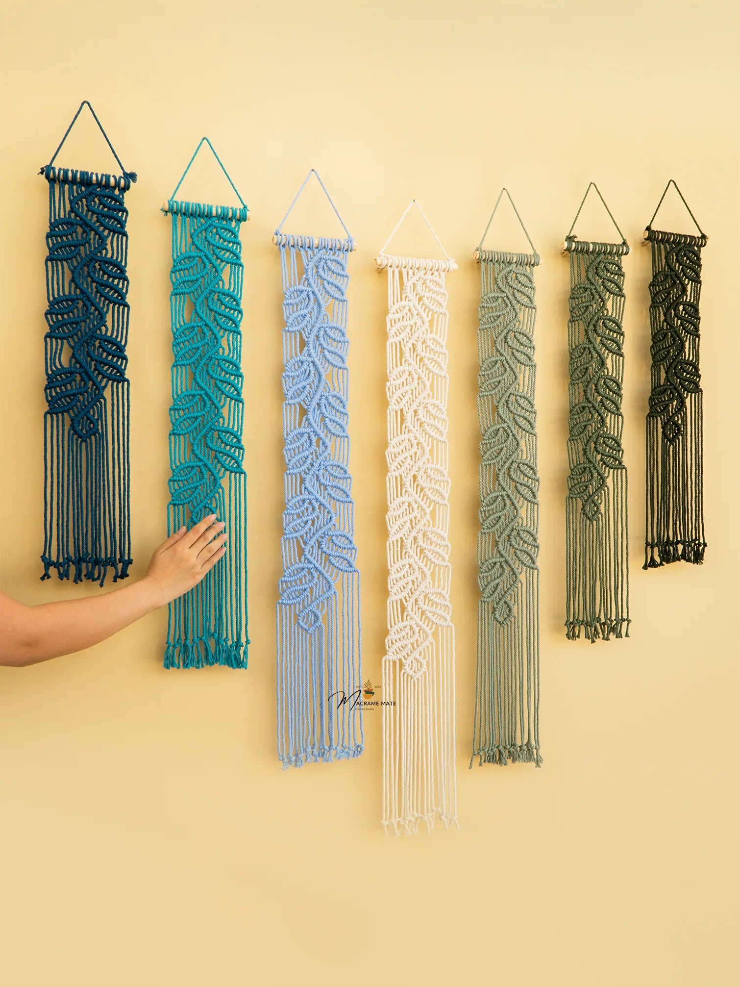 Set of six macrame wall hangings in various colors on a beige wall.