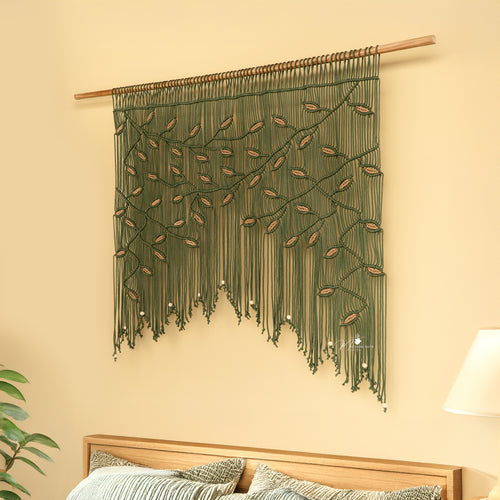 Macrame wall hanging with green and brown threads on a beige wall above a wooden bed.