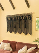 Black macrame wall hanging on a beige wall above a brown sofa with decorative pillows.