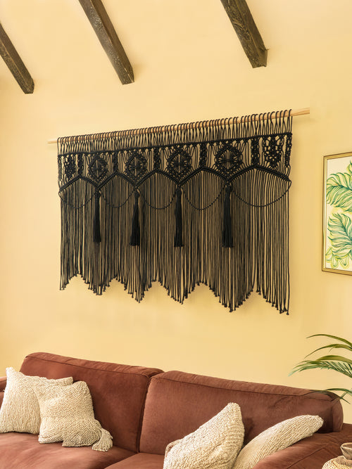 Black macrame wall hanging on a beige wall above a brown sofa with decorative pillows.