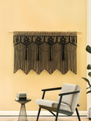 Decorative black macrame wall hanging on a yellow wall with a chair and side table in the foreground.