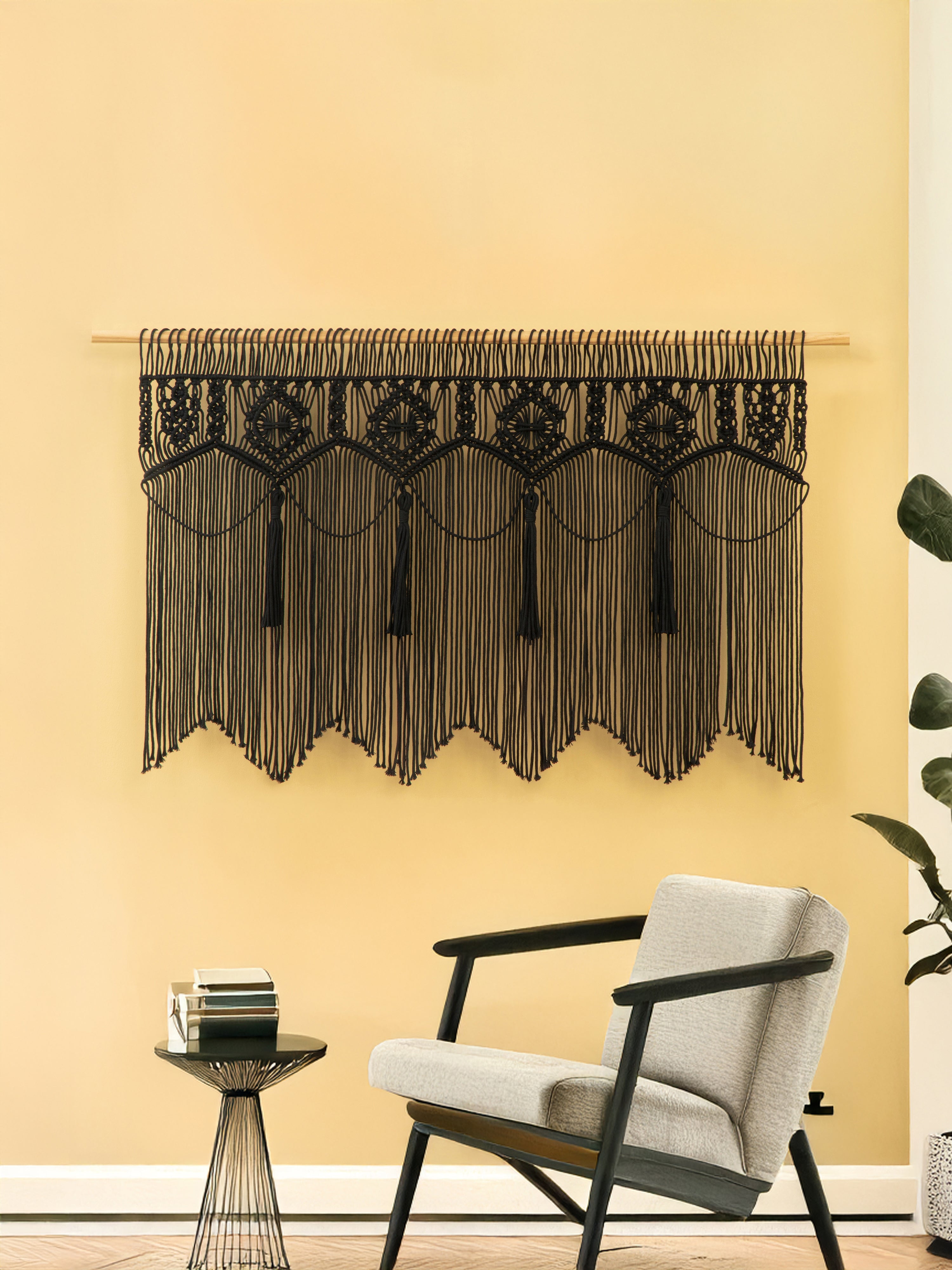 Decorative black macrame wall hanging on a yellow wall with a chair and side table in the foreground.