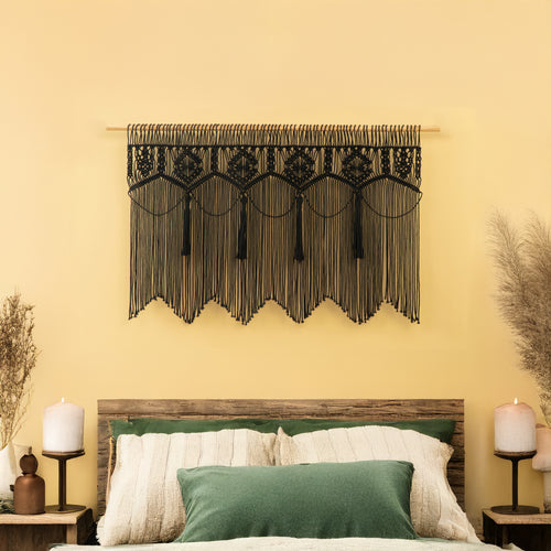 Decorative wall hanging with tassels above a bed with green and white pillows on a yellow wall.