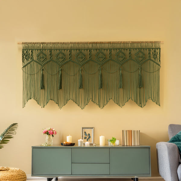 Decorative wall hanging with tassels above a bed with green and white pillows on a yellow wall.