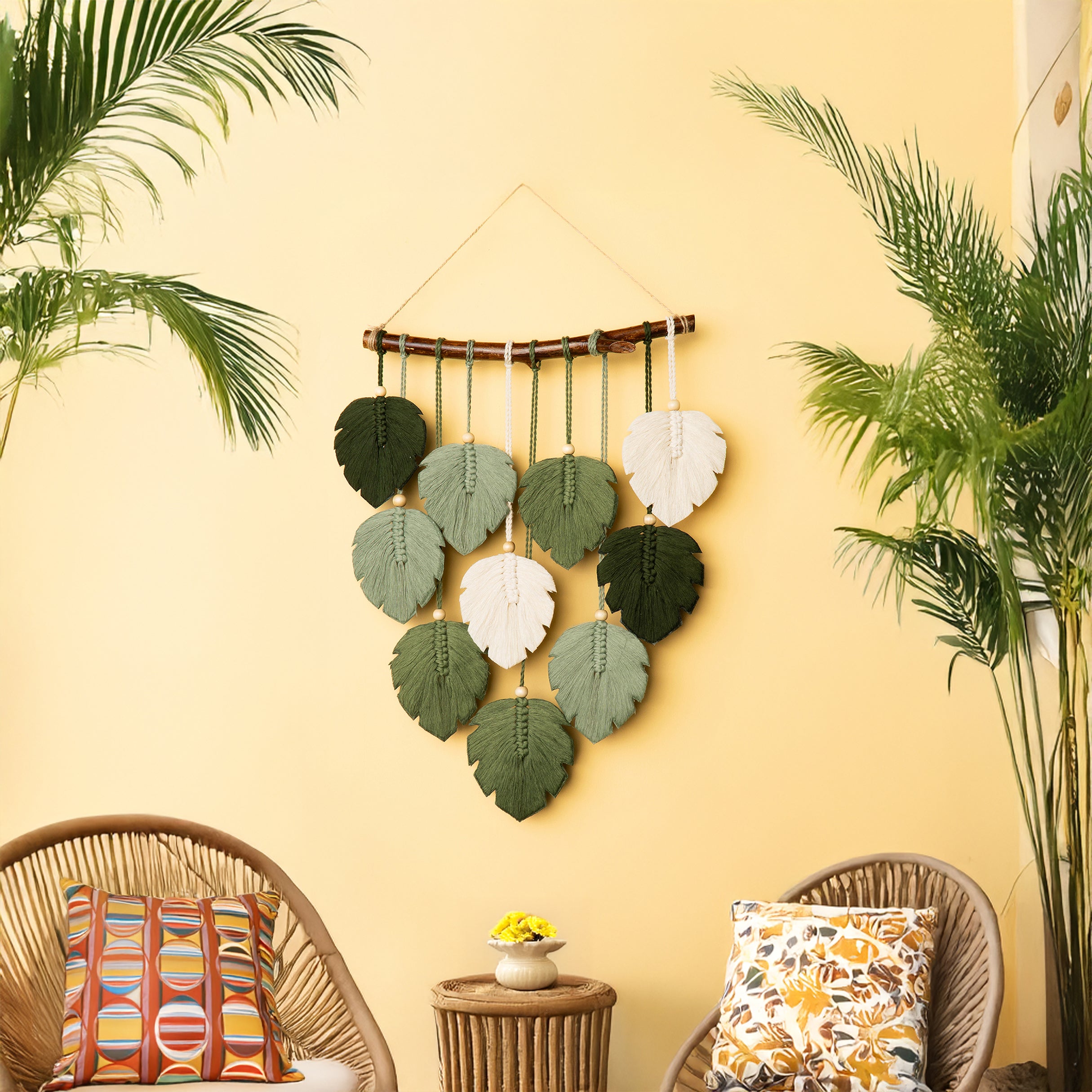Decorative wall hanging with leaf patterns on a yellow wall, with two wicker chairs and plants in the foreground.