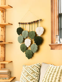 Decorative wall hanging with leaf-shaped objects on a yellow wall next to a wooden shelf.