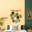 Decorative wall hanging with leaf patterns on a yellow wall, with two wicker chairs and plants in the foreground.
