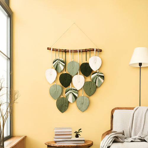 Decorative wall hanging with leaf patterns on a yellow wall in a room with a lamp and books.