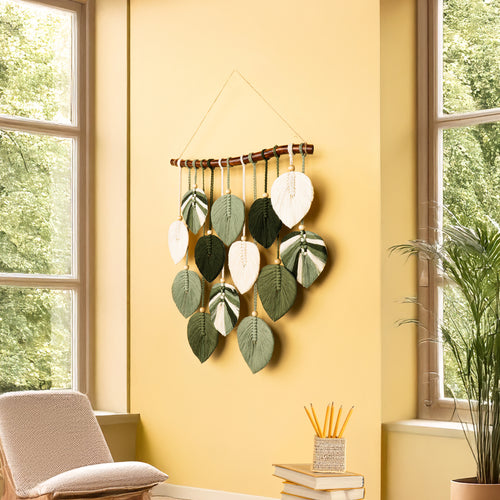 Decorative wall hanging with leaf patterns on a yellow wall in a room with windows and plants.