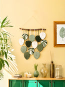 Decorative wall hanging with leaf patterns on a yellow wall, with a green cabinet and plants in the foreground.