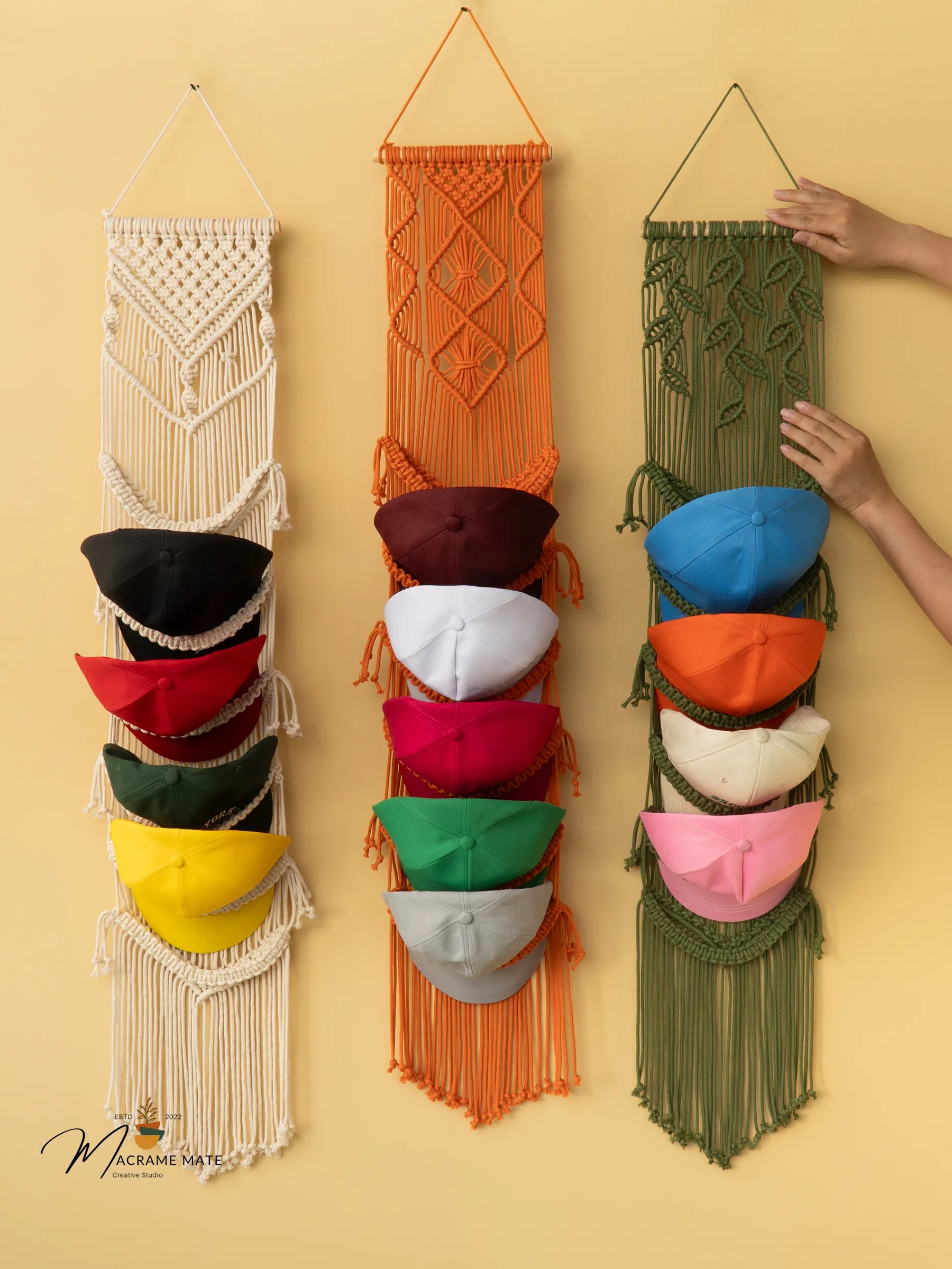 Set of four macrame hat racks in different colors with hats on a yellow background.