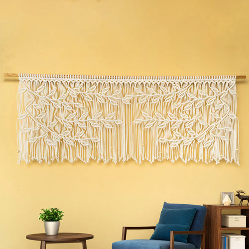 Macrame wall hanging with leaf patterns on a yellow wall, with a blue chair and small table in the foreground.