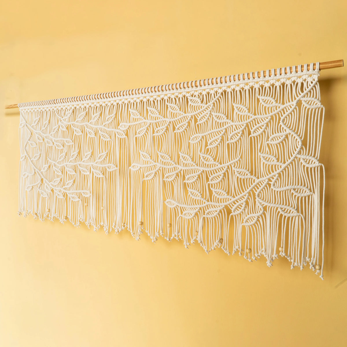 A white macrame wall hanging with an intricate leaf pattern, suspended from a light wood dowel against a solid pale yellow wall.