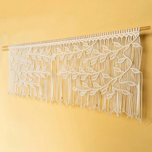 A white macrame wall hanging with an intricate leaf pattern, suspended from a light wood dowel against a solid pale yellow wall.