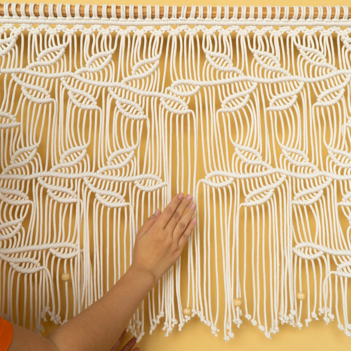 A close-up of a white macrame wall hanging with a leaf pattern, suspended from a wooden dowel. A person's hand is shown touching the hanging to demonstrate its texture and scale against a warm yellow background.