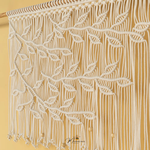 A close-up of a woven wall hanging or textile panel with an orange-brown braided rope pattern forming leaf-like shapes over a warm tan background, with small round white hanging beads visible.