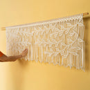 A white macrame wall hanging with an intricate leaf pattern suspended from a wooden rod, shown against a warm yellow wall with a person's arm touching it.