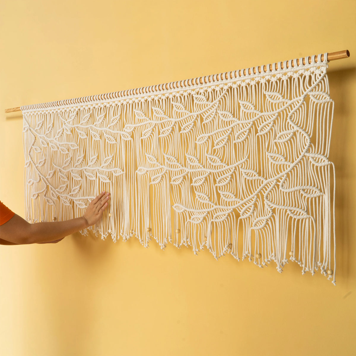 A white macrame wall hanging with an intricate leaf pattern suspended from a wooden rod, shown against a warm yellow wall with a person's arm touching it.
