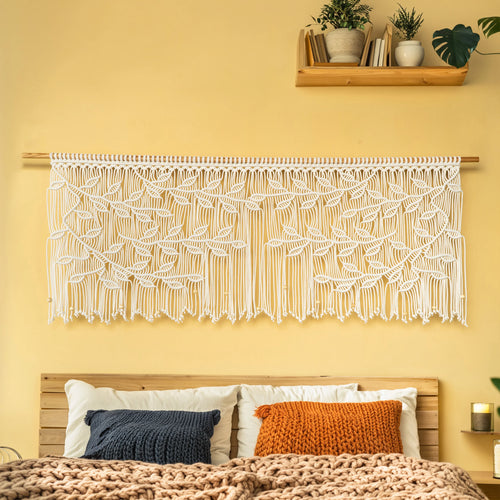 A large white macrame wall hanging with intricate leaf patterns is displayed on a wooden rod above a bed with a yellow wall and a wooden headboard.