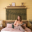 A woman in a beige sweater hangs a large woven macrame wall hanging with leaf patterns on a yellow paneled wall in a sunlit room.
