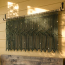 A cream macrame wall hanging with leafy vine patterns and hanging fringe, mounted on a wooden rod against a warm beige wall above a wooden console table.