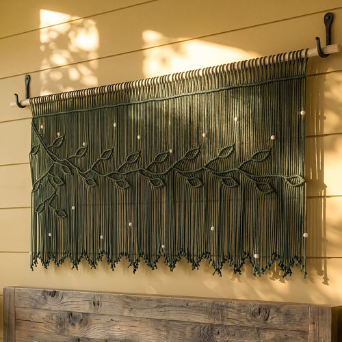 A cream macrame wall hanging with leafy vine patterns and hanging fringe, mounted on a wooden rod against a warm beige wall above a wooden console table.