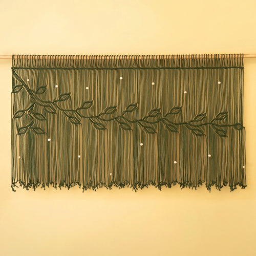 A dark green macrame wall hanging with a leafy vine design and small white bead accents, displayed against a warm beige wall.