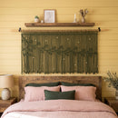 A cream macrame wall hanging with a leafy vine pattern suspended from a wooden rod above a bed in a bedroom with yellow paneled walls.