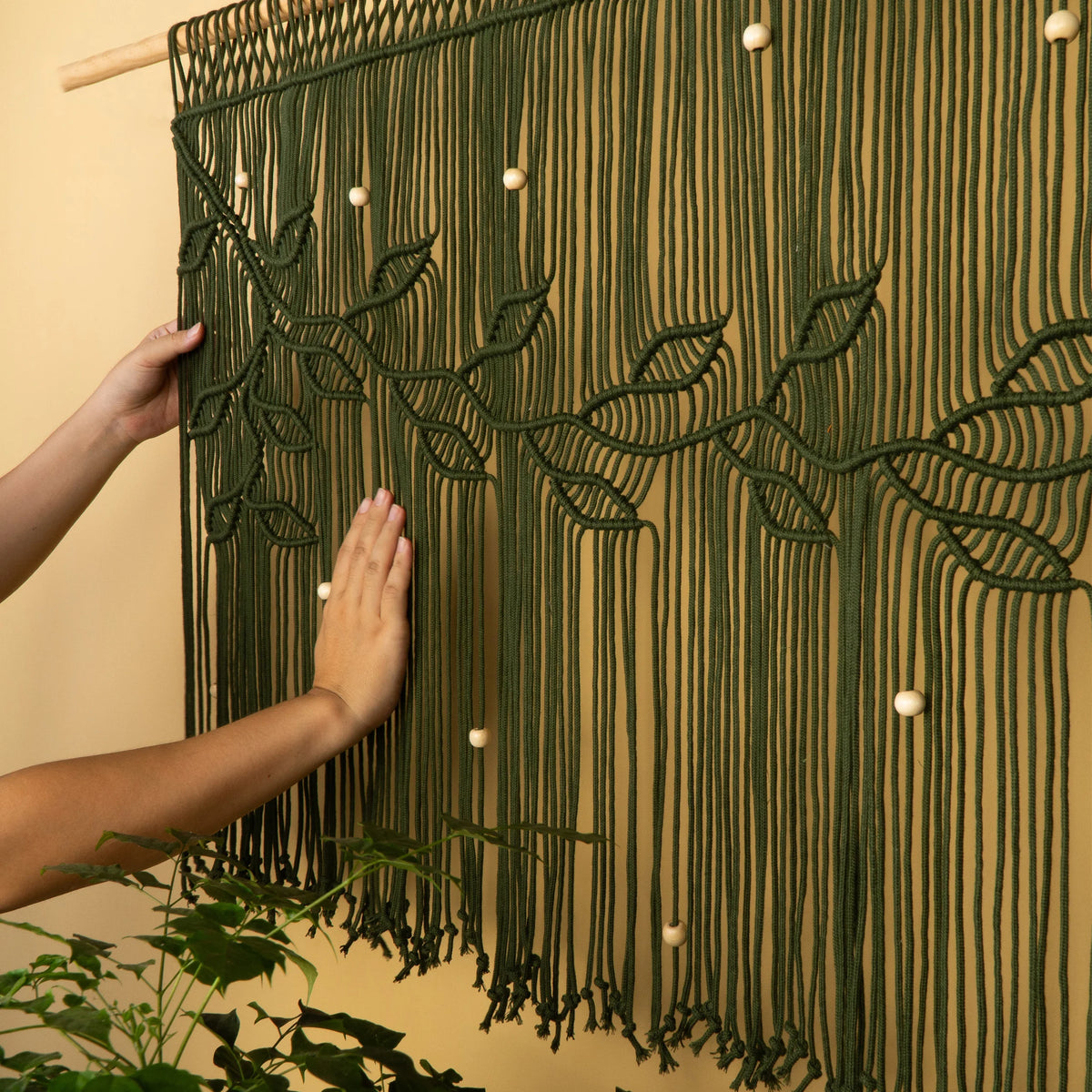 A cream-colored macrame wall hanging with intricate leaf patterns and long fringe, shown being held by two hands against a warm beige wall with a green plant in the foreground.
