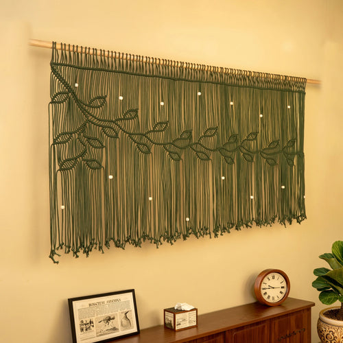 An olive green macrame wall hanging with a leafy vine pattern and small white bead accents, suspended from a wooden rod on a beige wall above a wooden console table with a clock and framed picture.