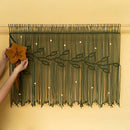 A dark green macrame wall hanging with a floral vine design is displayed on a warm beige wall. A person's hands are visible on the left, holding a large mustard-yellow fabric flower to demonstrate how to hang or style the piece.