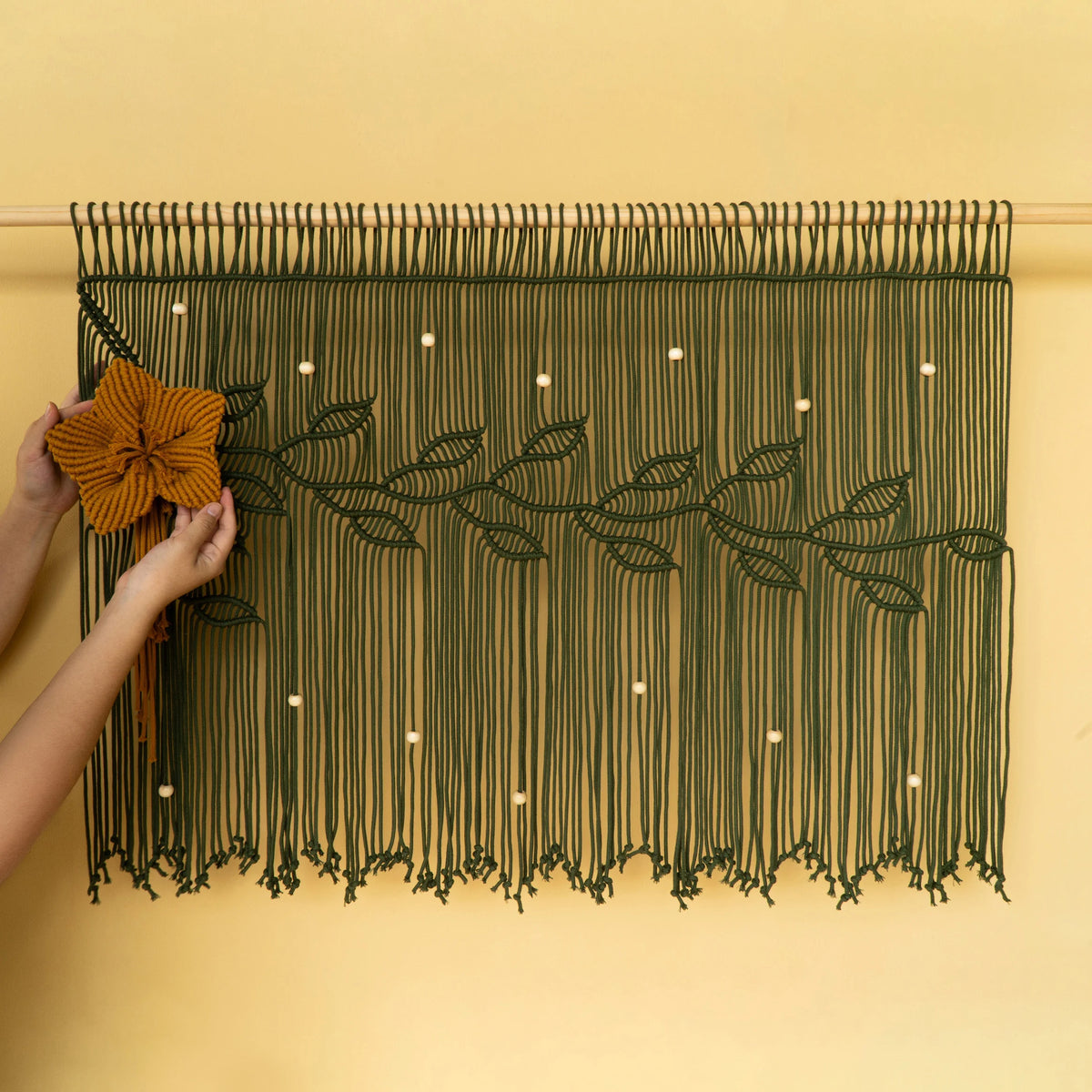 A dark green macrame wall hanging with a floral vine design is displayed on a warm beige wall. A person's hands are visible on the left, holding a large mustard-yellow fabric flower to demonstrate how to hang or style the piece.