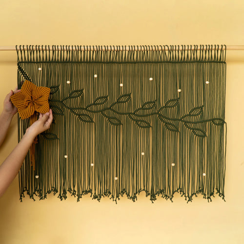 A dark green macrame wall hanging with a floral vine design is displayed on a warm beige wall. A person's hands are visible on the left, holding a large mustard-yellow fabric flower to demonstrate how to hang or style the piece.