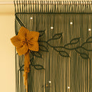 A close-up of a woven macrame wall hanging with a large mustard yellow flower and tassel on the left, dark green leafy vine patterns across the surface, and small white bead accents against a warm cream background.
