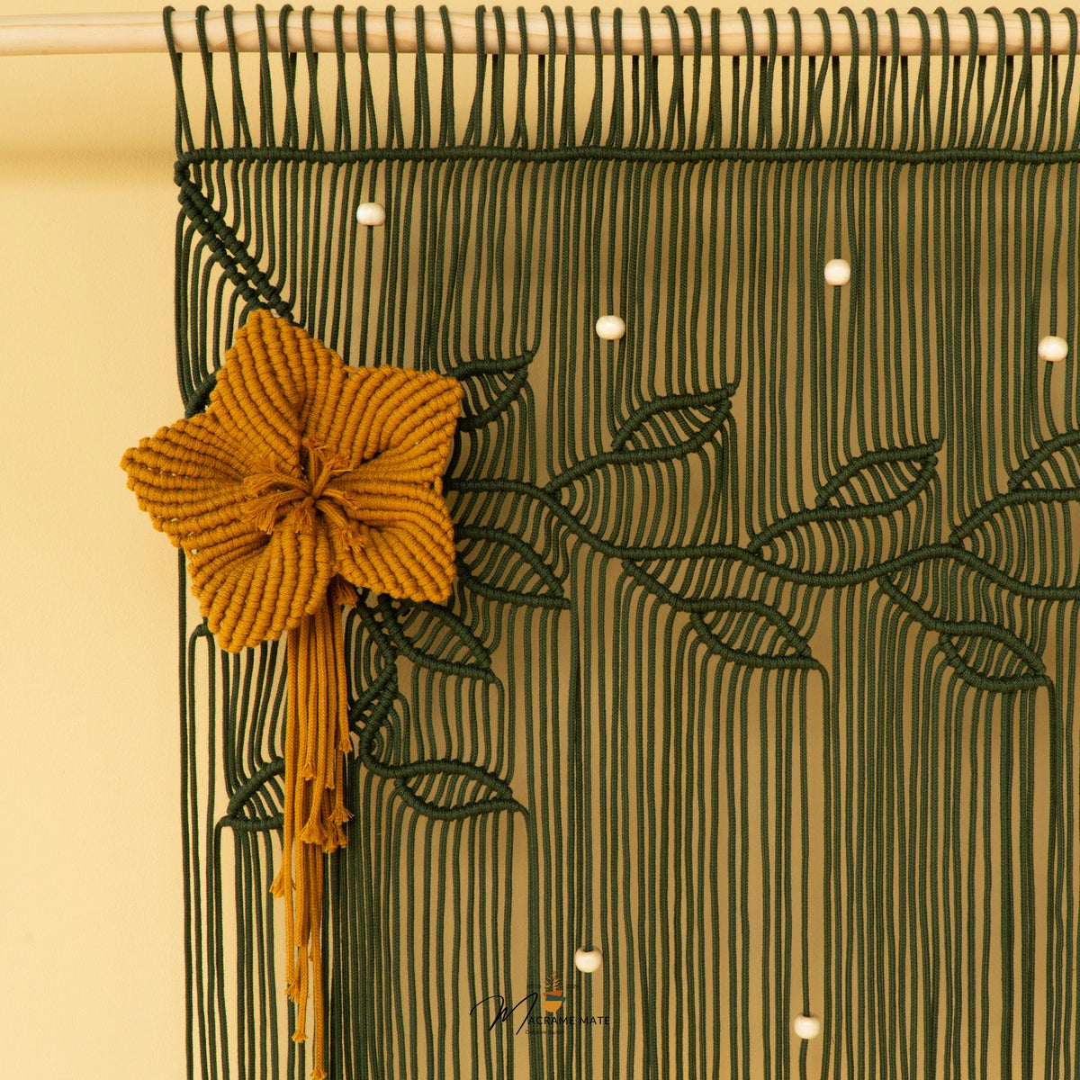 A close-up of a woven macrame wall hanging with a large mustard yellow flower and tassel on the left, dark green leafy vine patterns across the surface, and small white bead accents against a warm cream background.