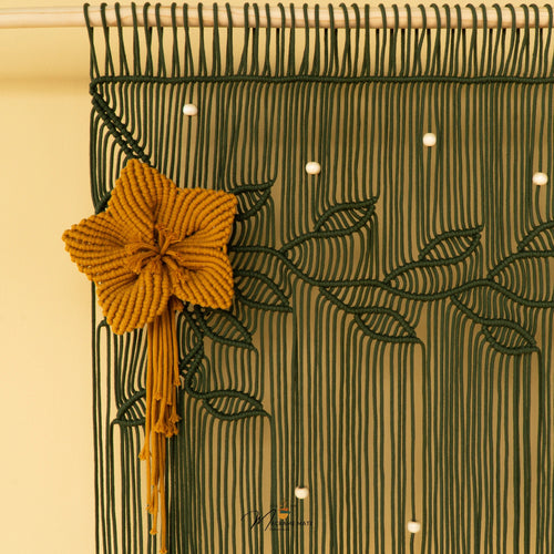 A close-up of a woven macrame wall hanging with a large mustard yellow flower and tassel on the left, dark green leafy vine patterns across the surface, and small white bead accents against a warm cream background.