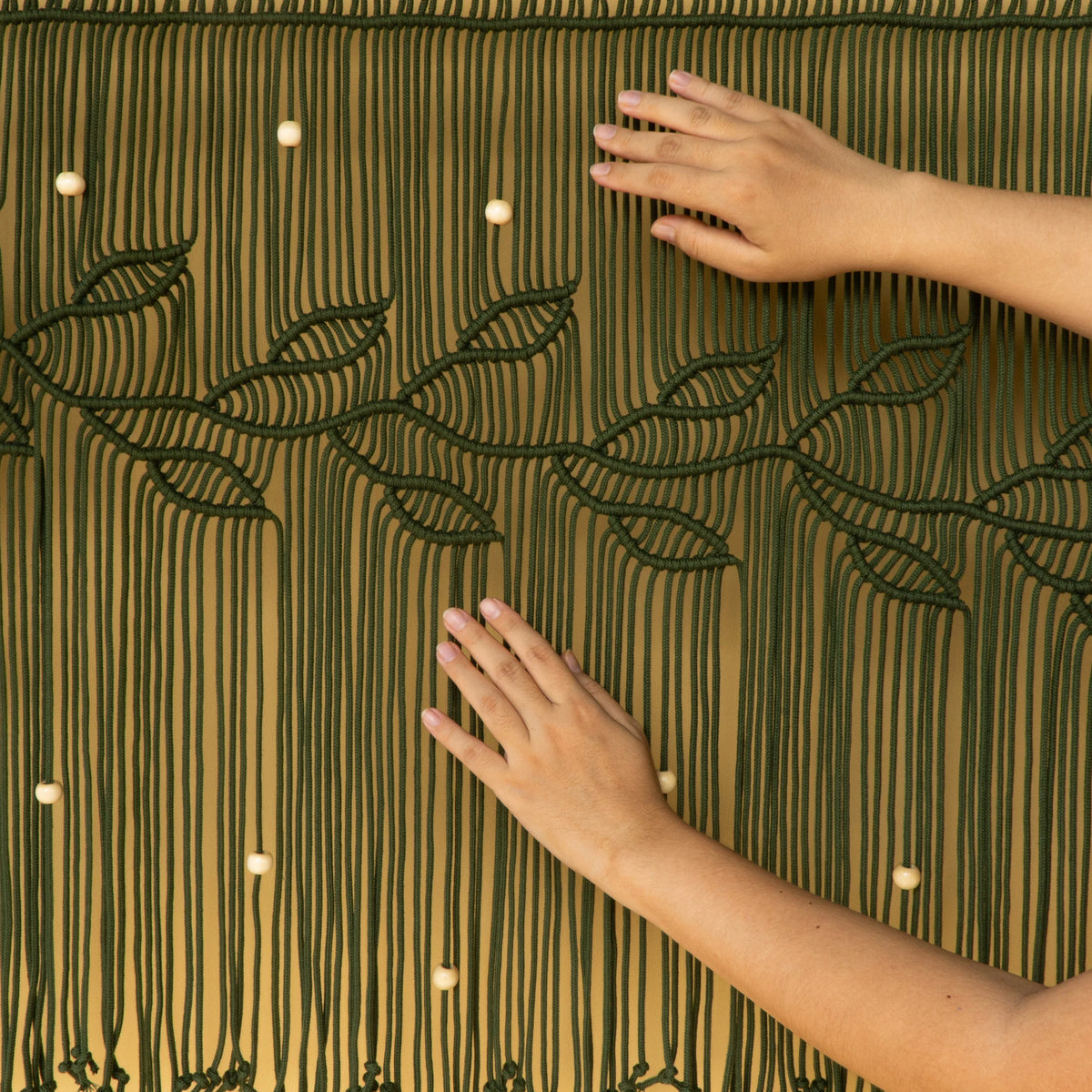 A dark green macrame wall hanging with leaf and vine patterns, suspended from a wooden rod. Two hands are shown adjusting the hanging, with a beige wall and a green plant visible in the background.