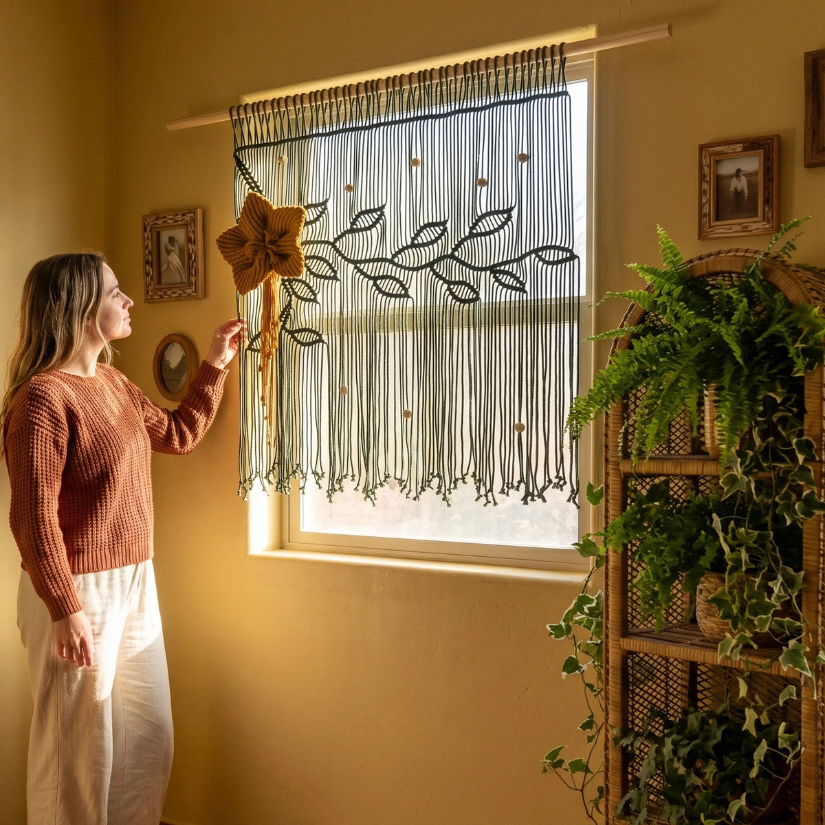 A woman in a rust-colored sweater and white pants stands by a window in a warmly lit room, holding a decorative brown curtain rod finial. The window is dressed with a sheer white curtain featuring a dark embroidered leaf pattern and long fringe tassels.