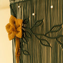 A close-up of a decorative macrame wall hanging with a dark green woven floral pattern, a prominent mustard yellow flower accent, and small white bead details against a warm beige background.