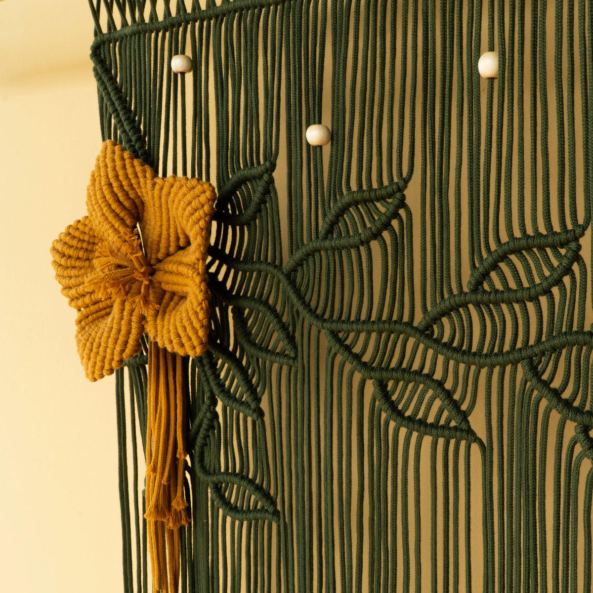 A close-up of a decorative macrame wall hanging with a dark green woven floral pattern, a prominent mustard yellow flower accent, and small white bead details against a warm beige background.