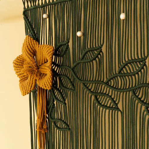 A close-up of a decorative macrame wall hanging with a dark green woven floral pattern, a prominent mustard yellow flower accent, and small white bead details against a warm beige background.