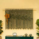 A dark olive green macrame wall hanging with a leafy vine design and a large mustard-yellow flower accent, displayed on a cream wall above a dark green console table with plants and decor.