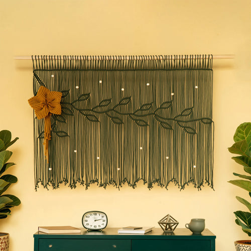 A dark olive green macrame wall hanging with a leafy vine design and a large mustard-yellow flower accent, displayed on a cream wall above a dark green console table with plants and decor.