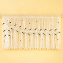 A white macrame wall hanging with woven leaf and vine motifs, displayed against a solid pale yellow background.