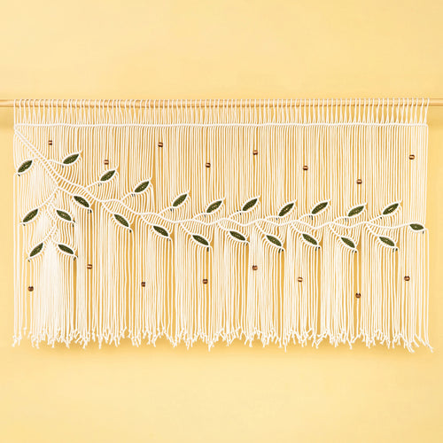 A white macrame wall hanging with woven leaf and vine motifs, displayed against a solid pale yellow background.