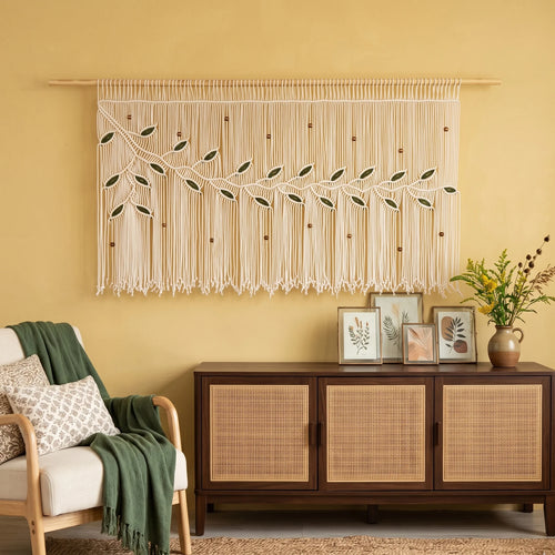 A decorative window curtain panel hangs in a window above a green sofa in a warm yellow living room. The curtain has a dark gray leafy vine design with a large yellow flower accent on the left side and long fringe at the bottom.