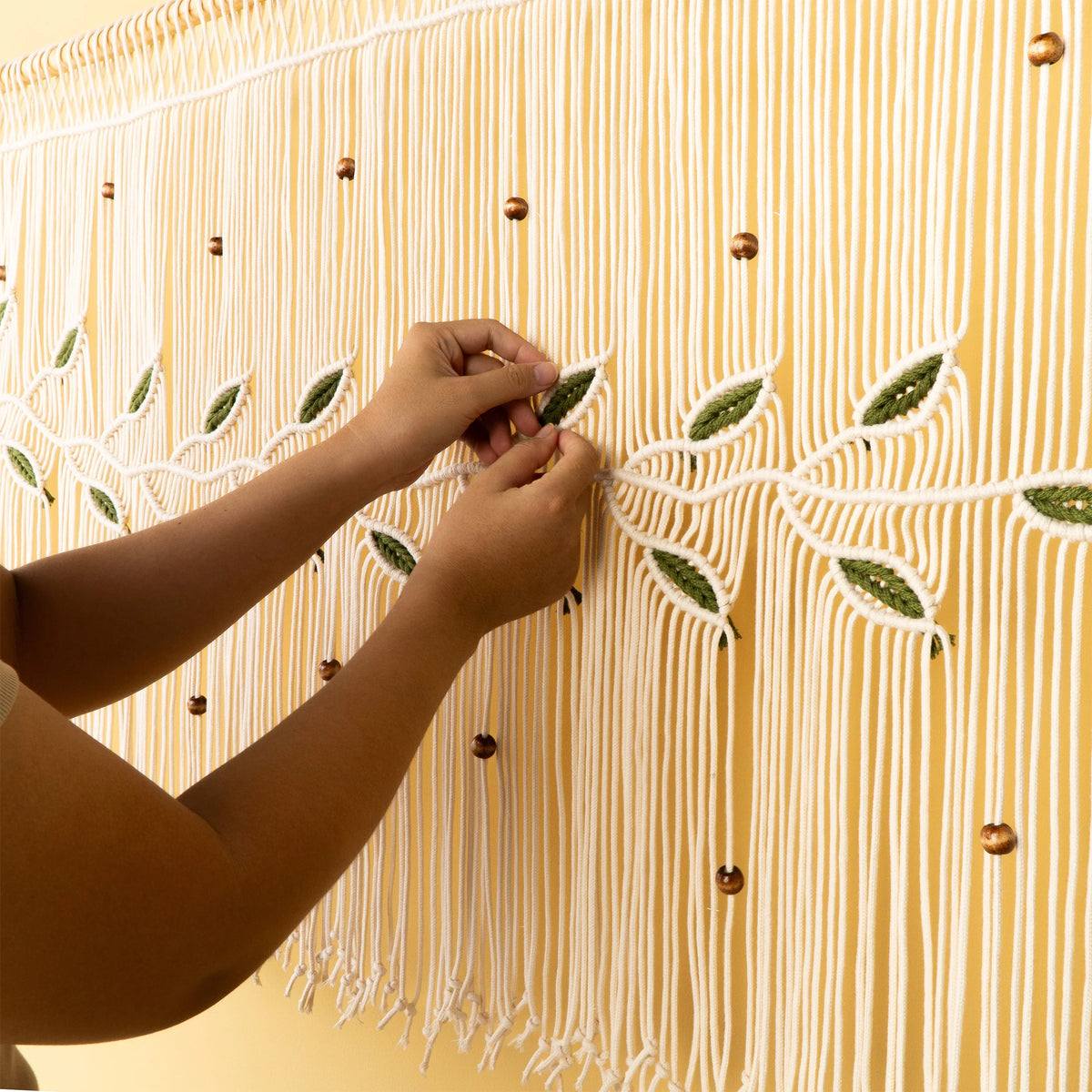 A person's hands are shown attaching a small green and white embroidered leaf to a large wall hanging or tapestry with white fringe, gold beads, and vertical stripes on a yellow background.