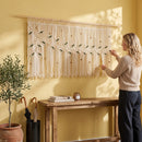 A woman sits on a bed in a cozy bedroom setting, holding an open book. The room features a large olive green macrame wall hanging with leaf patterns, styled with pink bedding, a wooden headboard, and a yellow shiplap wall.